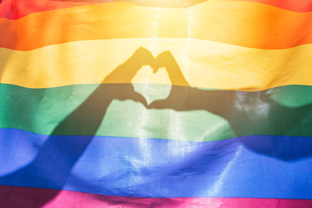 LGBTQ σημαία χρώματα ουράνιο τόξο, ένα σύμβολο για την ομοφυλοφιλική κοινότητα, δύο χέρια κάνοντας ένα σχήμα καρδιάς λάμπει από το φως του ήλιου. Gay σημαία για τον μήνα υπερηφάνειας και την ημέρα εορτασμού της σεξουαλικής και ερωτικής πολυμορφίας - Φωτογραφία, εικόνα