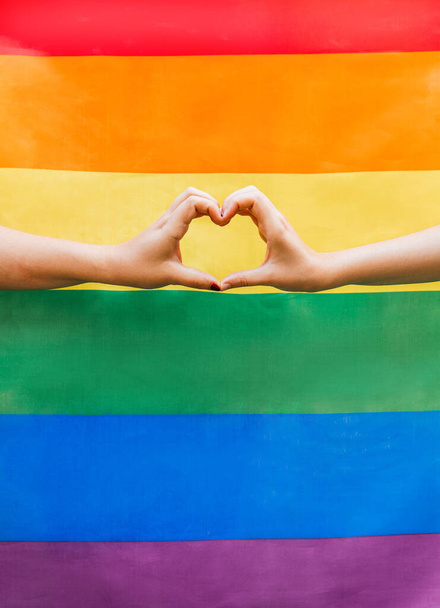 Закройте руки двух женщин, образующих сердце с радужным флагом ЛГБТК на заднем плане. Символ для гомосексуального сообщества месяца гордости и празднования дня сексуального и любовного разнообразия - Фото, изображение