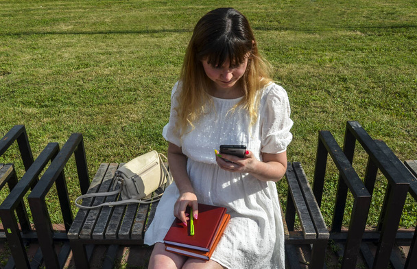 Okouzlující dívka s poznámkami nebo knihami sedí na lavičce v parku a dívá se do telefonu  - Fotografie, Obrázek