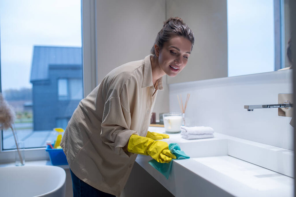A young woman in yellow gloves cleaning the surfaces in the bathroom - Fotó, kép