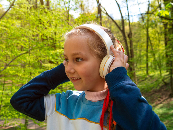 Adorable child girl in headphones learning language listening to music podcast with smartphone online outdoor in park. Stylish kid listens audio books tales Distance elementary school lesson in gadget - Foto, imagen