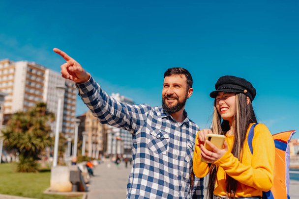 λευκός άνδρας δίνει οδηγίες σε μια νεαρή Ασιάτισσα τουρίστρια σε μια ευρωπαϊκή πόλη. Κινέζα που κάνει τουρισμό στην Γκιζόν, Αστούριες, Ισπανία - Φωτογραφία, εικόνα