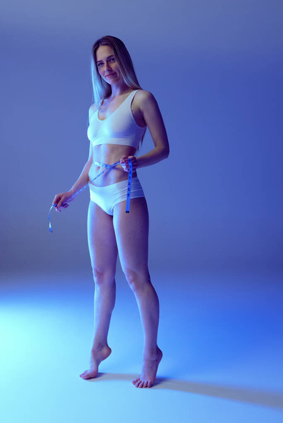 Full-length portrait of slim young woman measuring waist, posing in white underwear isolated over blue studio background in neon light - Foto, afbeelding