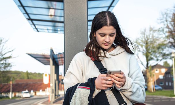 A young woman uses a smartphone while standing at a bus stop. - Foto, Bild