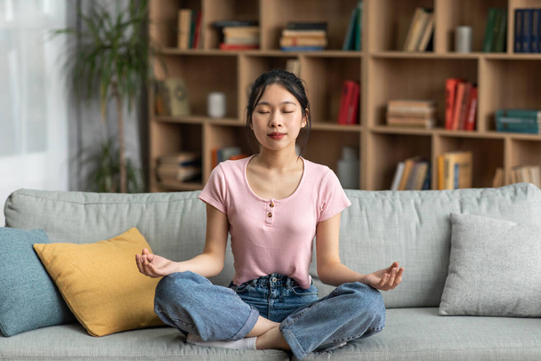 Rauhallinen aasialainen nainen istuu mukavalla sohvalla ja meditoi suljetuin silmin kotona, täyspitkä, kopioi tilaa - Valokuva, kuva
