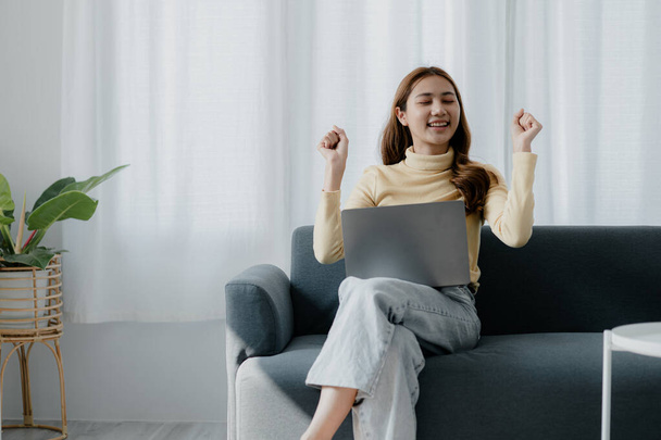 Asian woman dancing to the music playing from laptop on sofa in her home living room, weekend break, relaxation from hard work, drinking coffee. Take a vacation after a hard day's work. - Foto, Imagem