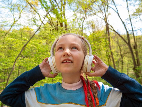 Kulaklıklı sevimli bir kız çocuğu parkta açık havada akıllı telefonuyla müzik podcast 'i dinliyor. Şık bir çocuk müzik kitapları dinler. Hikayeler, uzaktan ilkokul dersi. - Fotoğraf, Görsel