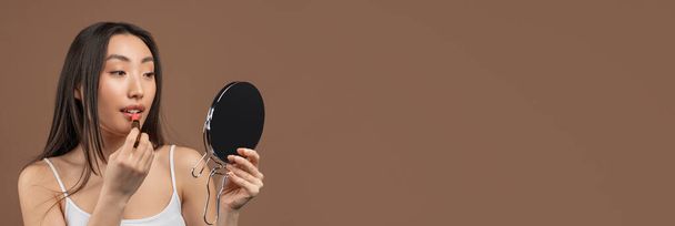 Pretty asian lady making daily makeup, using mirror and lipstick over brown studio background, panorama with free space - Foto, immagini