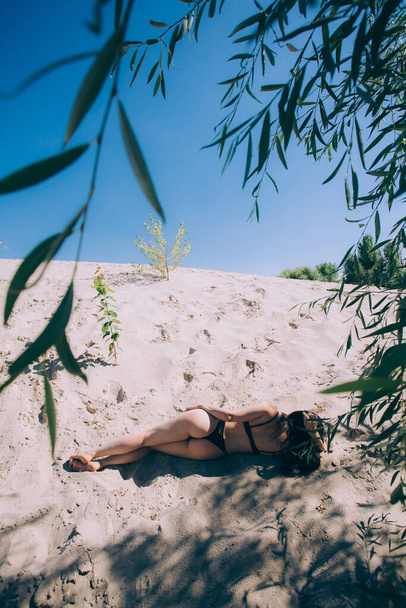 Une œuvre d'art. Une fille en maillot de bain noir se trouve sur le sable sous les arbres par une journée ensoleillée. - Photo, image