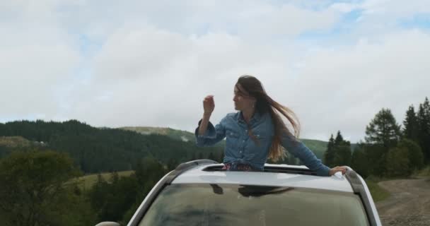 風の中で長い髪の毛がなびいています。女の子は彼女の髪をまっすぐにし、屋根を通って車の屋根の上の自然を見ています。昼間、緑の森、丘、雲。自然への車の旅. - 映像、動画