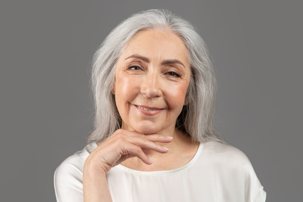 Krásný portrét starší ženy dotýkající se obličeje, dívající se do kamery a usmívající se na šedém pozadí studia - Fotografie, Obrázek