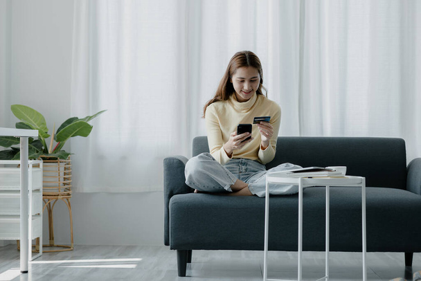 A woman playing smartphone on the sofa in her home living room, she is relaxing on weekends after a hard day's work, she is shopping online and paying by credit card. Credit card payment concept. - Fotoğraf, Görsel
