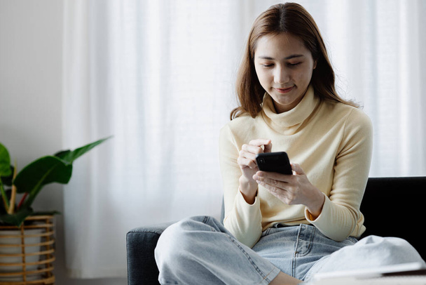 A woman playing smartphone on the sofa in her home living room, she is relaxing on weekends after a hard day's work, she is shopping online and paying by credit card. Credit card payment concept. - Foto, immagini