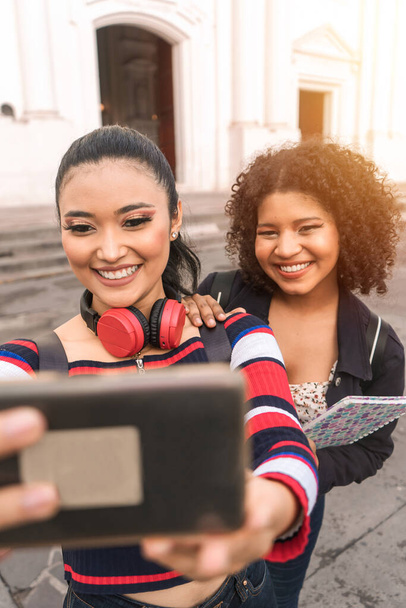 Vertical photo of two multiracial Latina students taking a self-portrait with their cell phone smiling outdoors in a public square in Leon - Photo, Image