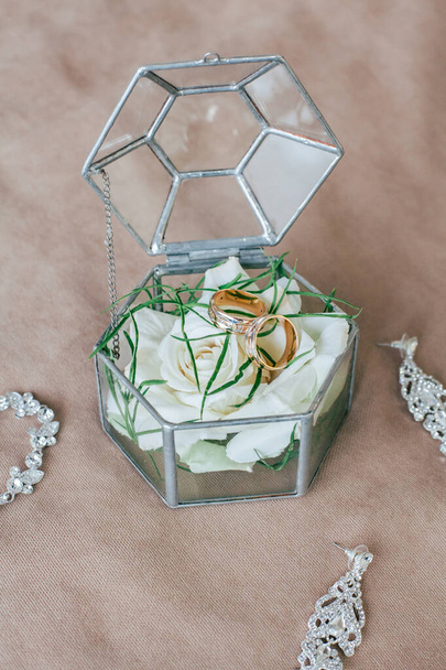 Декоративная стеклянная подарочная коробка для свадьбы, на бежевом фоне. Внутри золотые обручальные кольца для невесты и жениха, а также белая роза и зеленые листья. - Фото, изображение
