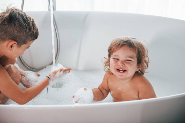 Two kids having fun and washing themselves in the bath at home. Posing for a camera. - 写真・画像