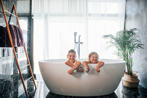 Two kids having fun and washing themselves in the bath at home. Posing for a camera. - 写真・画像