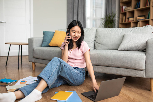 Happy korean lady talking on smartphone and looking at laptop screen, sitting on floor near sofa at home, copy space - Photo, Image