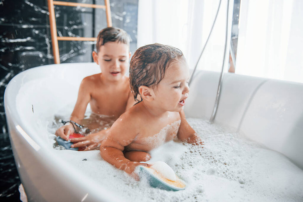 Two kids having fun and washing themselves in the bath at home. Helping each other. - Photo, image