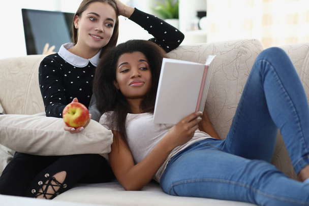 Mejores amigos leyendo libro escalofriante en sofá, manzana para merienda, ambiente relajante - Foto, Imagen