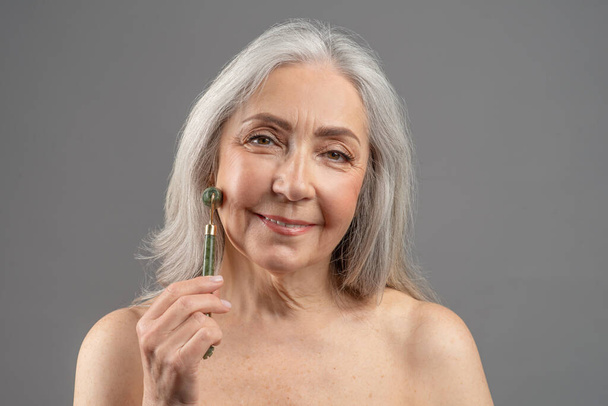 Charmante Seniorin macht Lymphdrainage Gesichts-Selbstmassage mit Jade-Rolle auf grauem Studiohintergrund - Foto, Bild