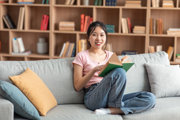 Domestic life concept. Positive asian lady holding paper book, sitting on comfy couch at home alone, free space - Photo, image