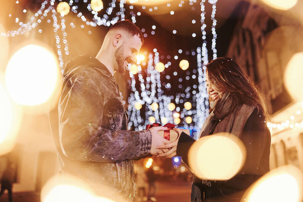 Holds gift box. Happy multiracial couple together outdoors in the city celebrating New year. - Foto, Imagem