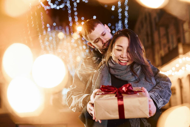 Holds gift box. Happy multiracial couple together outdoors in the city celebrating New year. - Foto, imagen