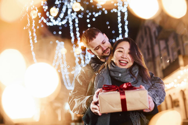 Holds gift box. Happy multiracial couple together outdoors in the city celebrating New year. - Valokuva, kuva