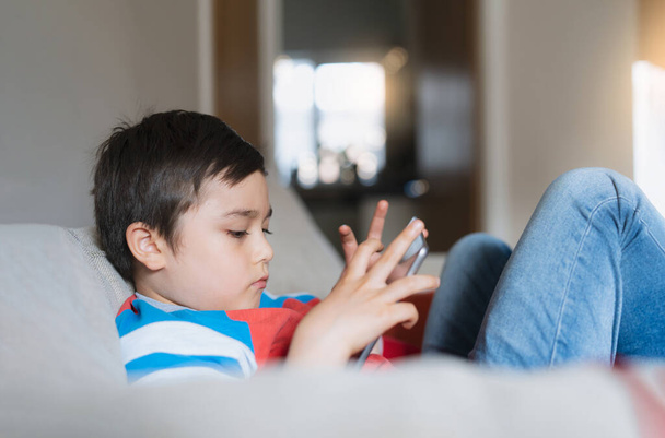 Jeune garçon utilisant une tablette de jeu sur Internet, Enfant assis sur le canapé regarder ou parler avec un ami en ligne, Enfant se détendre dans le salon le matin, Enfants avec une nouvelle technologie concept - Photo, image