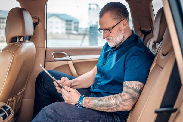 Σοβαρός ηλικιωμένος επιχειρηματίας χρησιμοποιώντας δισκίο στο εσωτερικό του αυτοκινήτου - Φωτογραφία, εικόνα