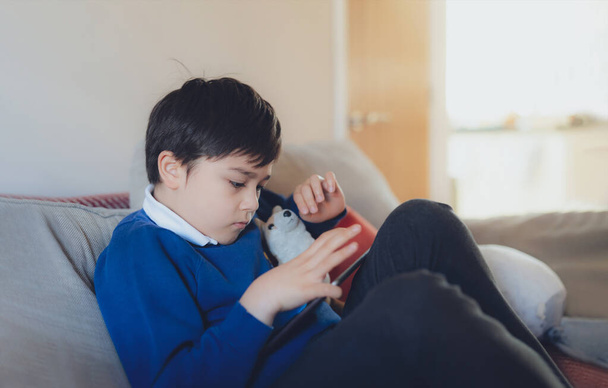 Happy schoolboy playing with dog toy and game online with friend on tablet,Kid using internet sending homework to the teacher, Positive child sitting on sofa relaxing in living room after school - Foto, Bild