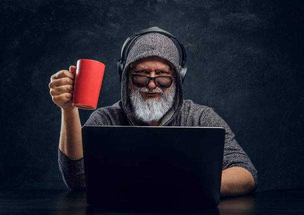 Stylish grandfather hacker with cup and headphones using laptop - Фото, изображение
