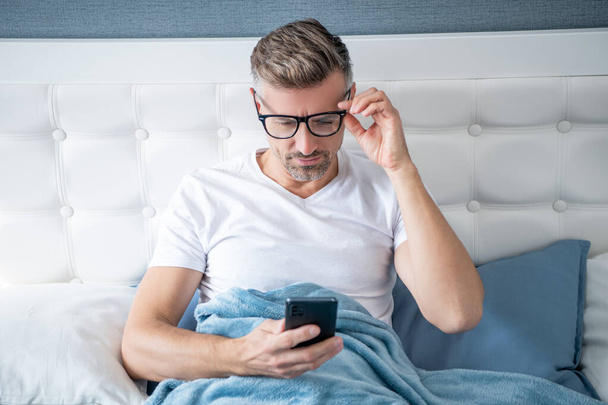 mature man in bed chatting on phone wearing eyeglasses - Foto, Imagen