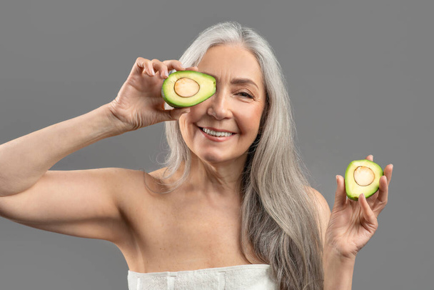 Органічні вітаміни для зрілої шкіри. Усміхнена старша жінка тримає розрізане авокадо перед очима на сірому фоні
 - Фото, зображення