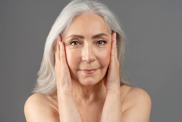 Portrait of feminine senior woman touching silky skin on her face, looking at camera over grey studio background - Photo, Image