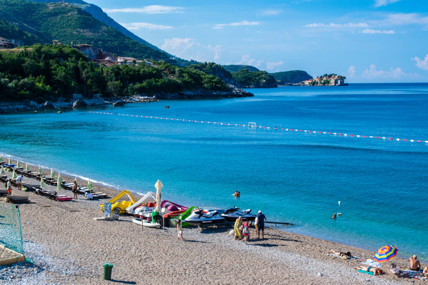 Rafailovici, Montenegro - July 3, 2021: Vacationers on Kamenovo pebble beach with sun loungers and umbrellas. Summer sunny morning. Blue sky. Sveti Stefan island backgound. Budva Riviera. - Photo, Image