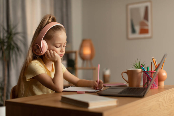 Tired Schoolgirl Learning Online Using Laptop And Taking Notes Indoor - Foto, imagen