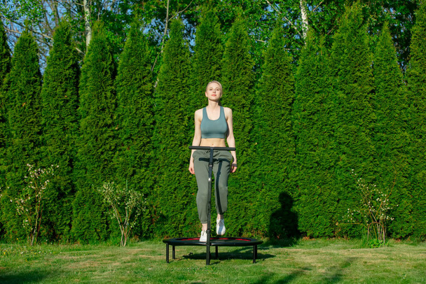 girl does Jumping fitness on trampoline in outdoor on green grass and trees on background - Fotografie, Obrázek