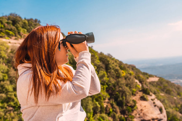 young woman using binoculars to spot birds in the sky, while hiking on top of mountain. person observing the horizon. - Фото, изображение