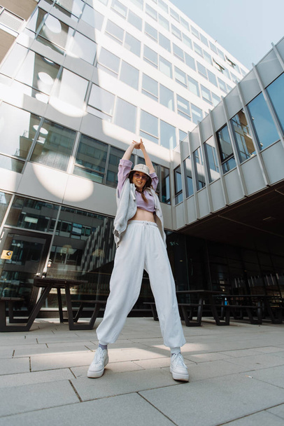 Fashion cutte woman with sunglasses and hat dancing outside looking at camera. Female dancer performing dance show making freestyle stunts. Funny stylish caucasian hip hopgirl. 4K - Fotó, kép