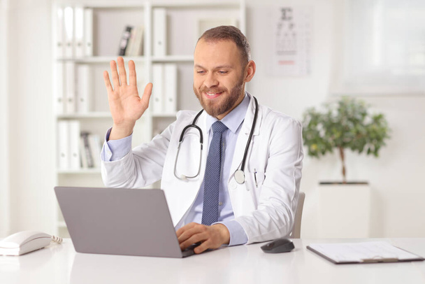 Male doctor sitting in an office and waving at a laptop computer - Φωτογραφία, εικόνα