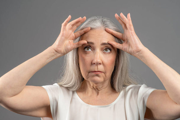 Portrait of senior woman dissatisfied with wrinkles on her forehead, touching face on grey studio background - Fotoğraf, Görsel