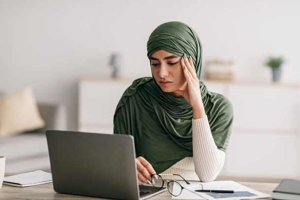 Несчастная молодая арабка-фрилансер в хиджабе устала работать на ноутбуке, страдает головной болью в домашнем офисе - Фото, изображение