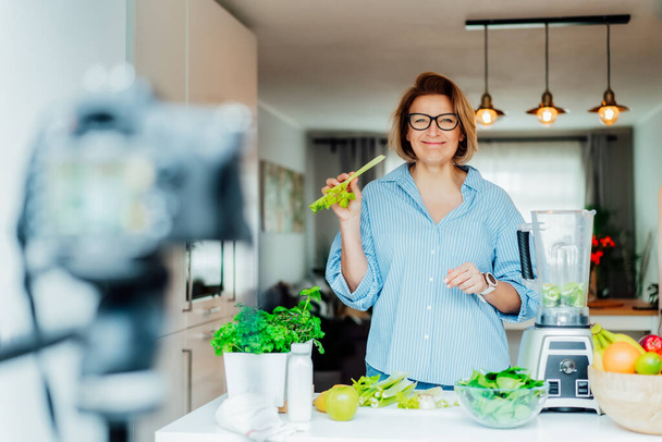 Middle age woman is blogging for her wellness lifestyle channel about healthy balanced living in the kitchen of her home. Creating video content for social media with camera on a tripod - Photo, image