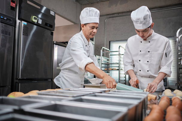 Two professional Asian male chefs in white cook uniforms and aprons are kneading pastry dough and eggs, preparing bread and fresh bakery food, baking in oven at stainless steel kitchen of restaurant. - Фото, изображение