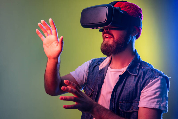 Impressed bearded hipster man standing in vr, playing video game and trying to touch something, wearing beanie hat and denim vest. Indoor studio shot isolated on colorful neon light background. - Photo, Image
