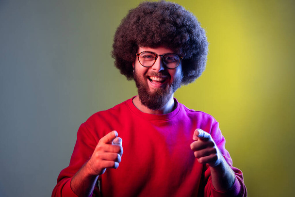 Portrait of hipster man with Afro hairstyle pointing to camera, smiling, making happy choice, we need you concept, wearing red sweatshirt. Indoor studio shot isolated on colorful neon light background - Foto, Bild