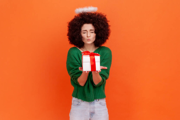 Positive funny angelic woman with Afro hairstyle wearing green casual style sweater giving wrapped present box, making kiss gesture, keeps eyes close. Indoor studio shot isolated on orange background. - Foto, Imagem