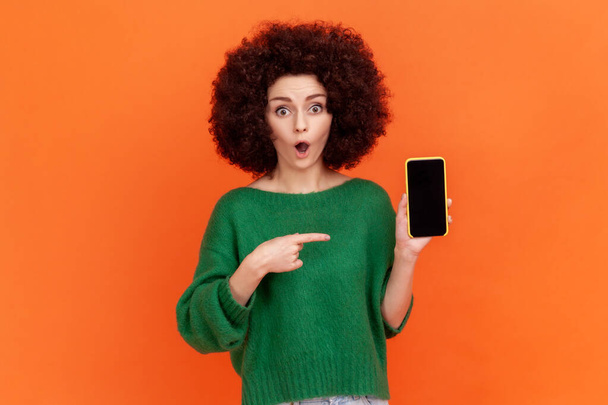 Astonished female woman with Afro hairstyle in green casual style sweater holding cell phone with empty display, presenting area for advertisement. Indoor studio shot isolated on orange background. - Photo, Image